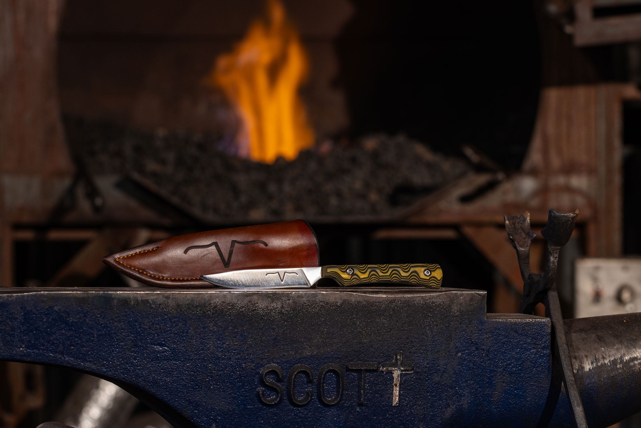 Flying V Bird Blade knife and leather sheath beside a forge, from the client’s product-page photography